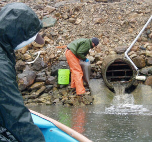 Two people in waterproof attire—one in foreground and one in background—standing near a rocky façade and stormwater drain taking water samples for stormwater pollution prevention planning.