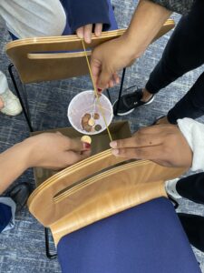 A plastic cup full of coins is surrounded by multiple hands holding uncooked spaghetti