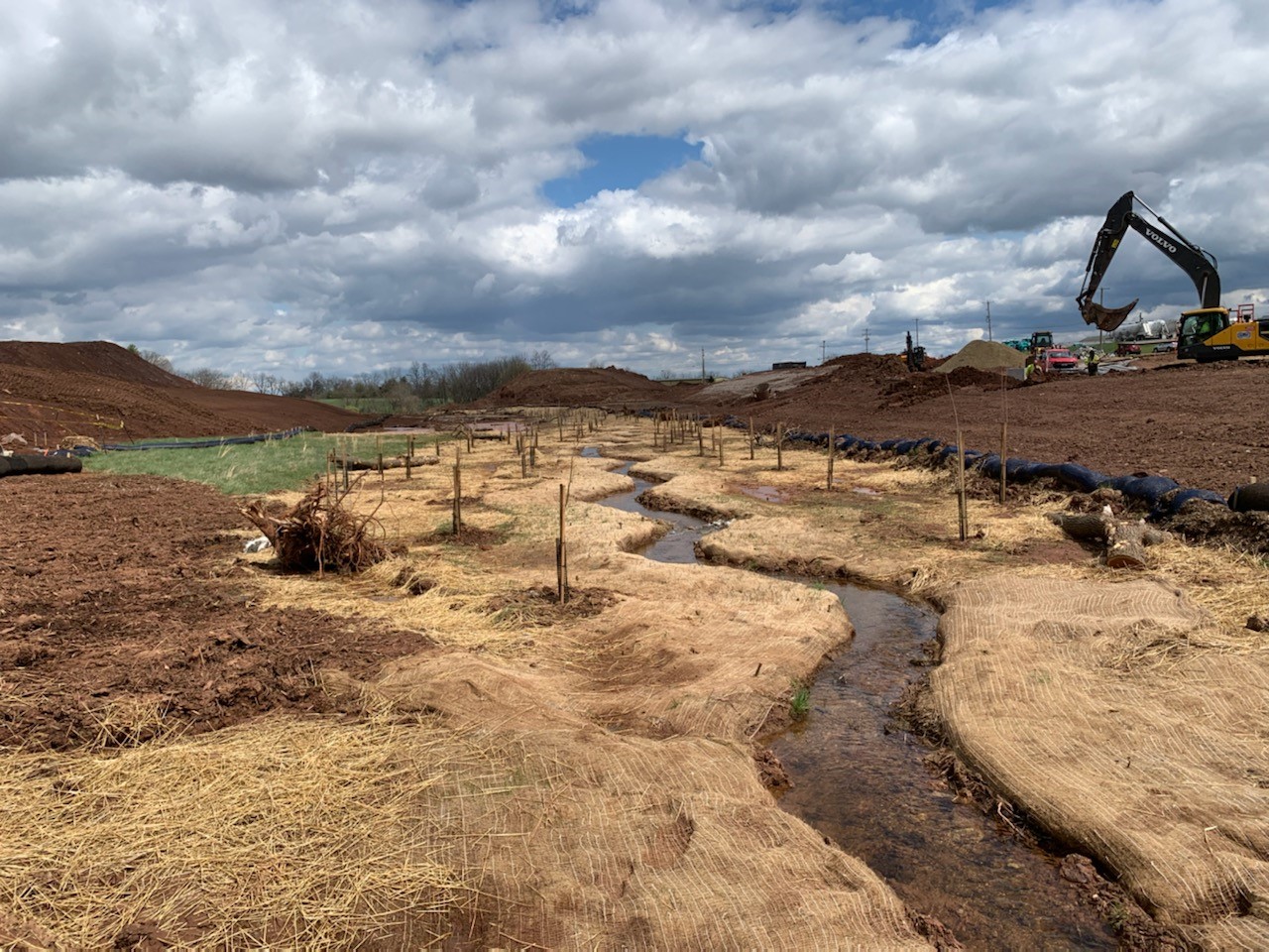Photo: foreground features a flat, grassy area with a narrow stream running through it. The grassy area is surrounded by gentle hills with excavation equipment sitting on the right side, all under a cloudy sky.