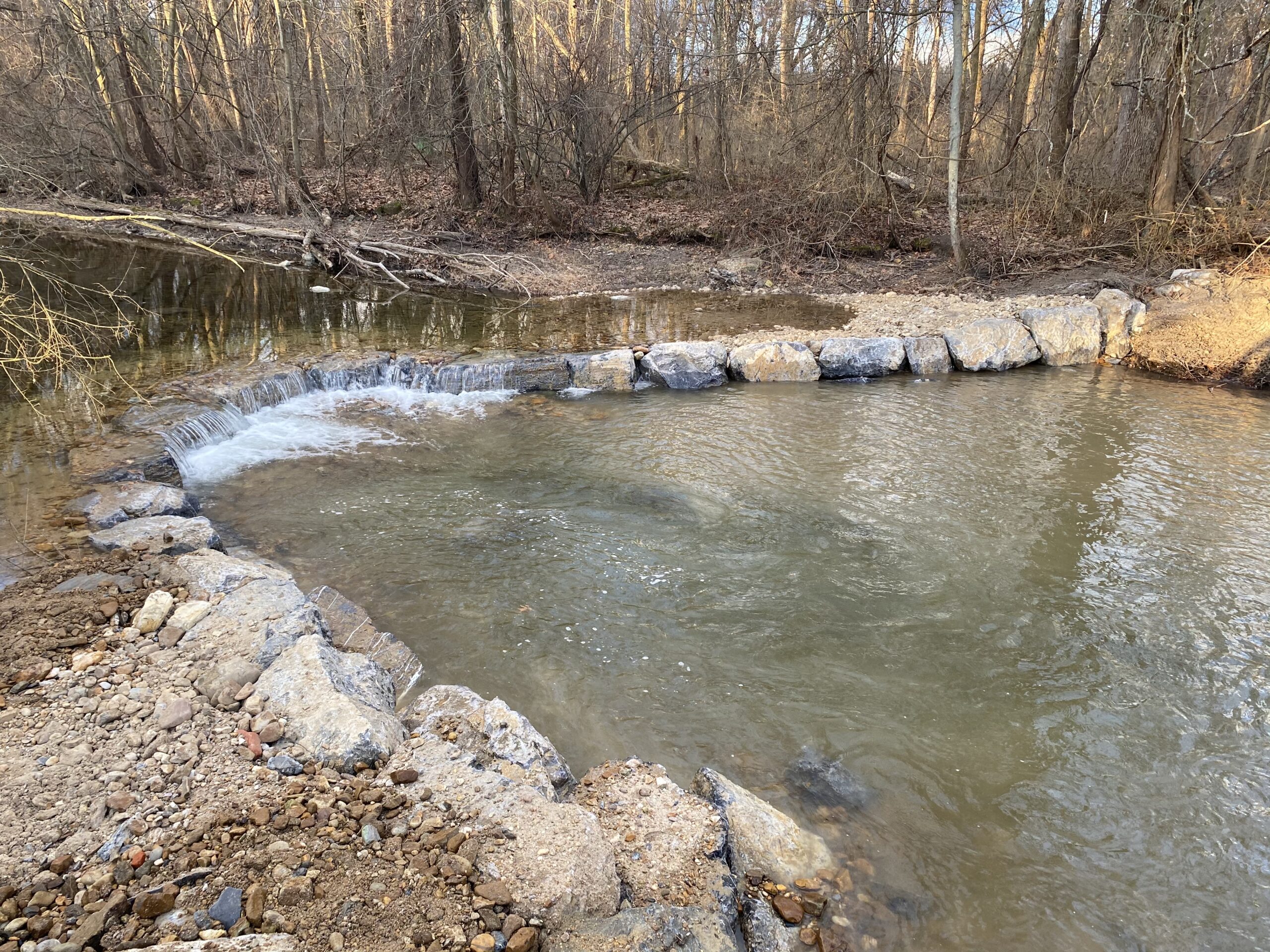 Photo: A small, clear stream flows through rocks that form a small dam. The surrounding area has leafless trees and a mix of rocky and muddy terrain, indicating late autumn or early winter.