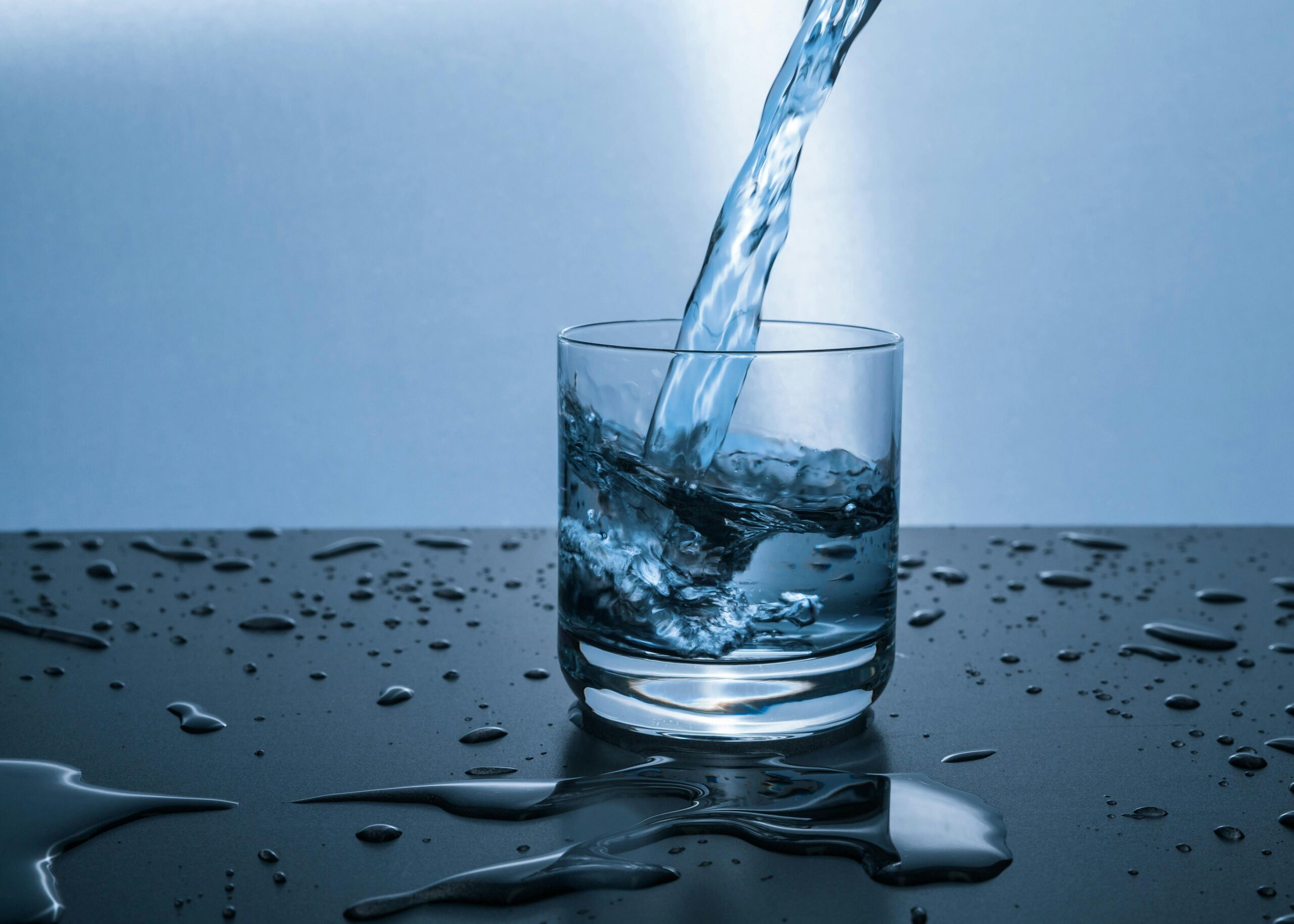 Photo: A short glass with water being poured into it from above. The glass is sitting on a black counter with water droplets on it.
