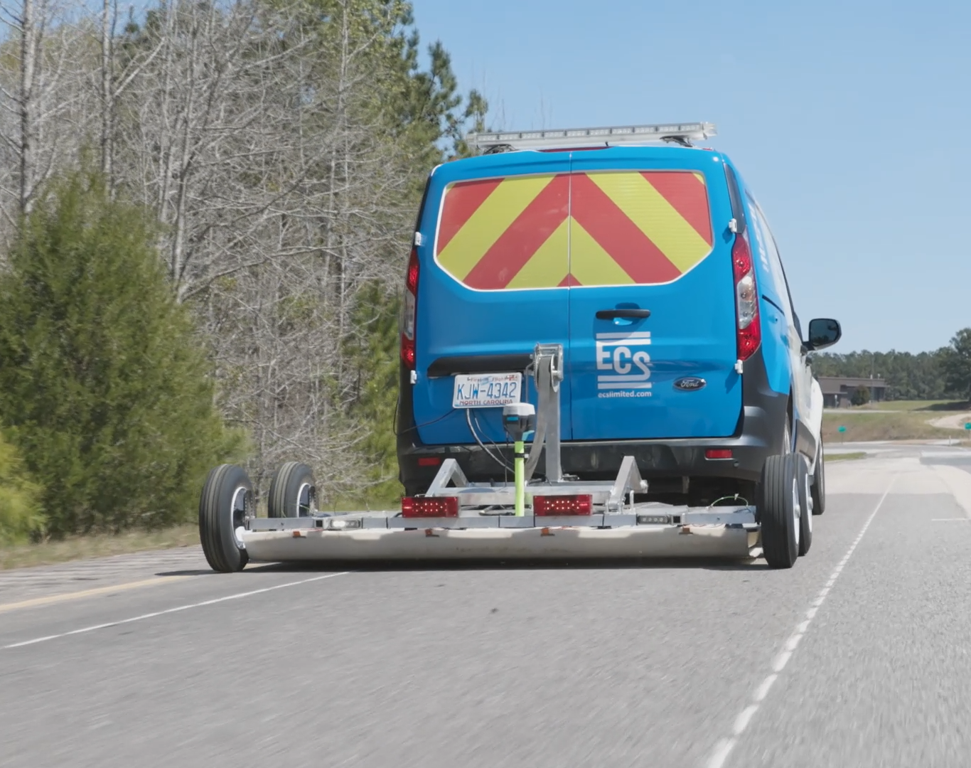 Photo of the back of a blue van with red and yellow diagonal stripes in the windows. The can is towing a white card low to the ground that is rigged with ground-penetrating radar technology.