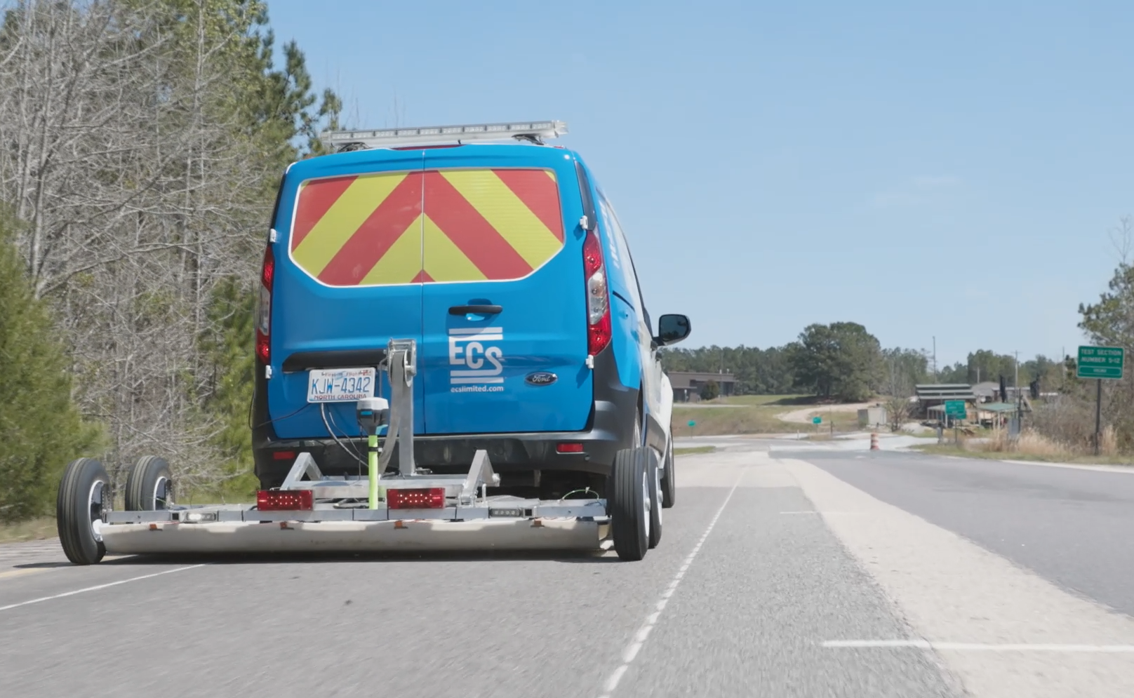 Photo of a blue and white van with red and yellow chevron on the back window towing a cart with ground-penetrating radar equipment on a road.