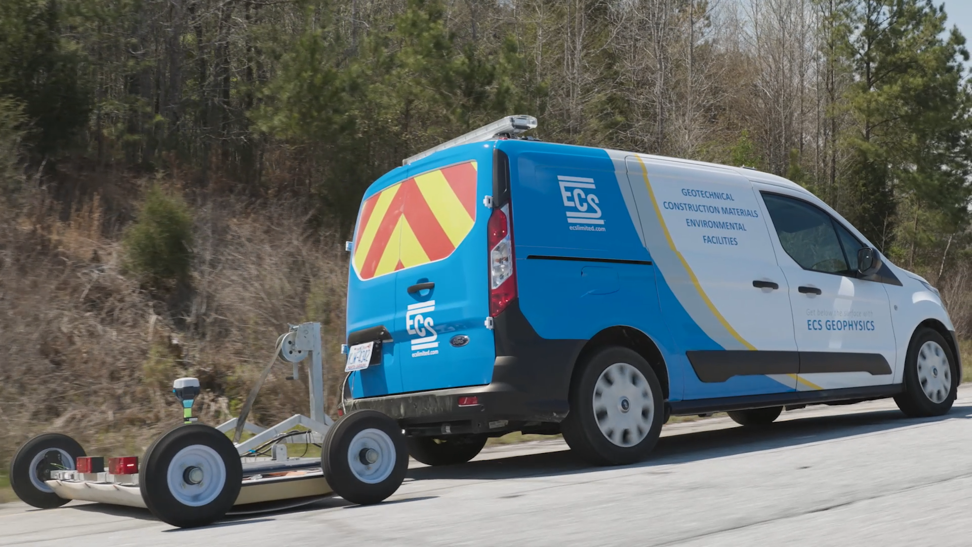 Photo of a blue and white van with red and yellow chevron on the back window towing a cart with ground-penetrating radar equipment on a road.