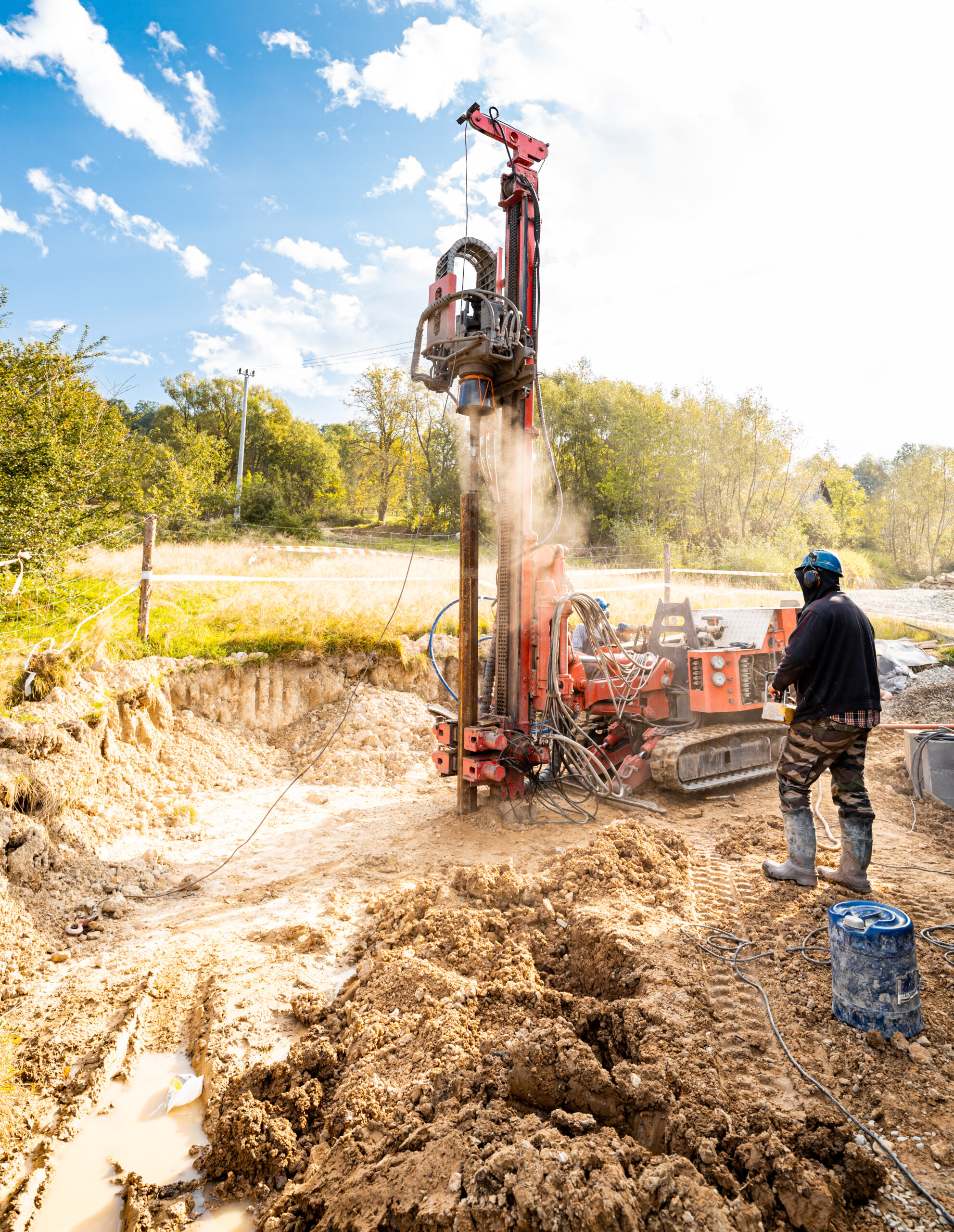 Photo of an orange drill drilling wells in the ground in a dirt work site. A worker in black clothes, a blue hard hat, and ear protect stands nearby