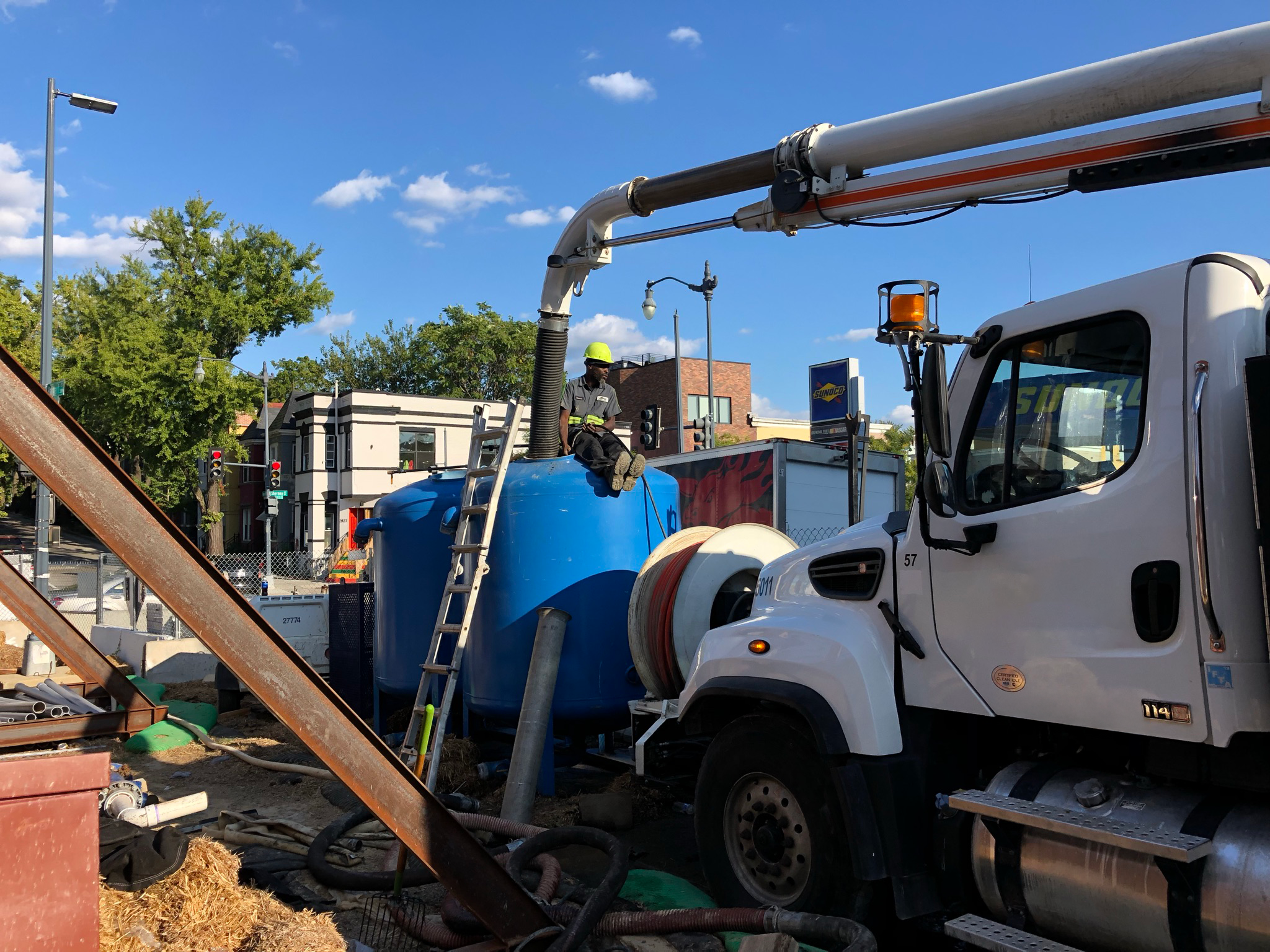Photo of a work site with a white truck with an overhead boom and hose leading into a large blue tank. A man in a neon yellow hard hat and grey outfit sits on the tank monitoring the process.