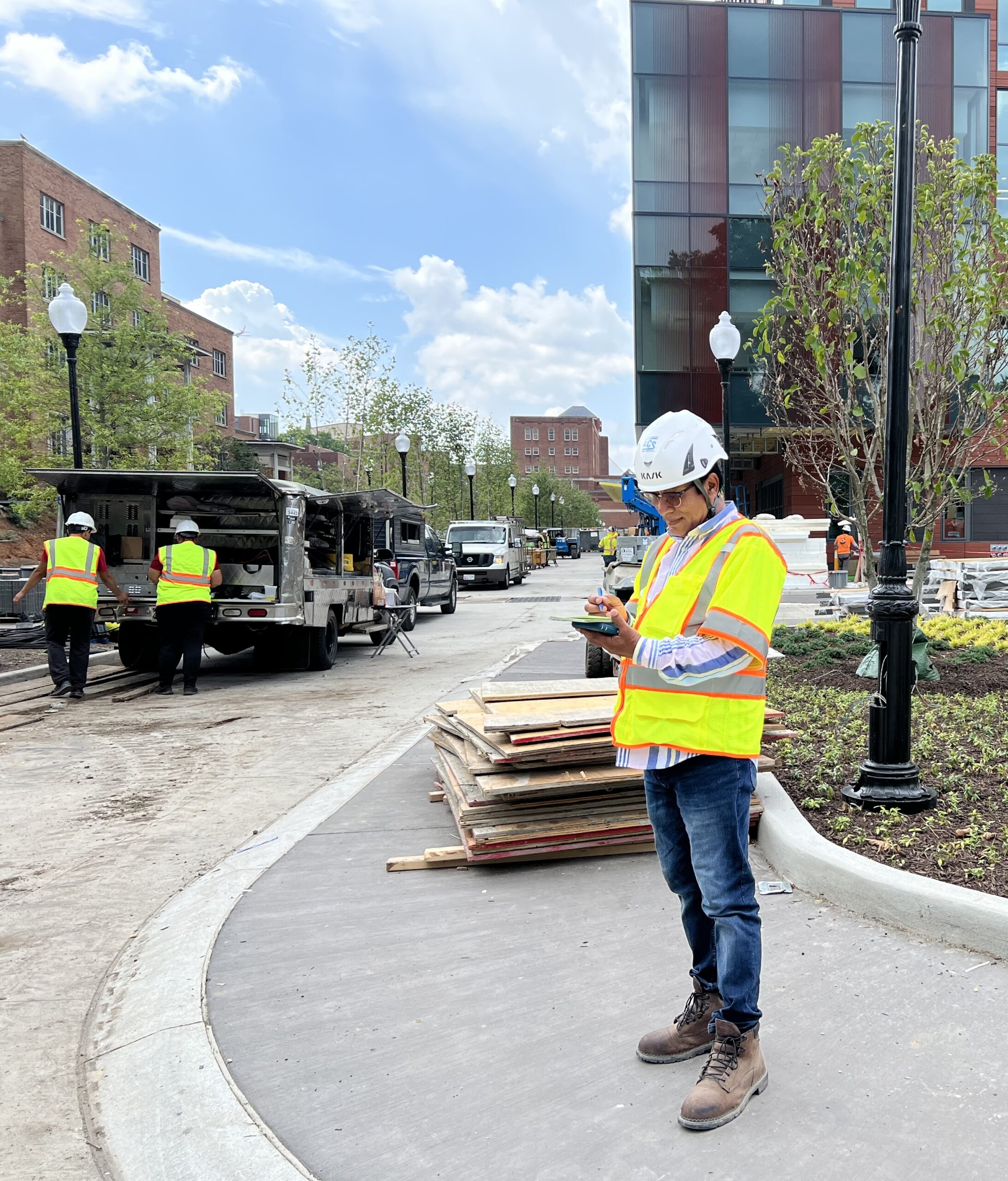 A man in a white hard hat and neon safety vest stands in front of buildings and a truck marking something on a notebook.