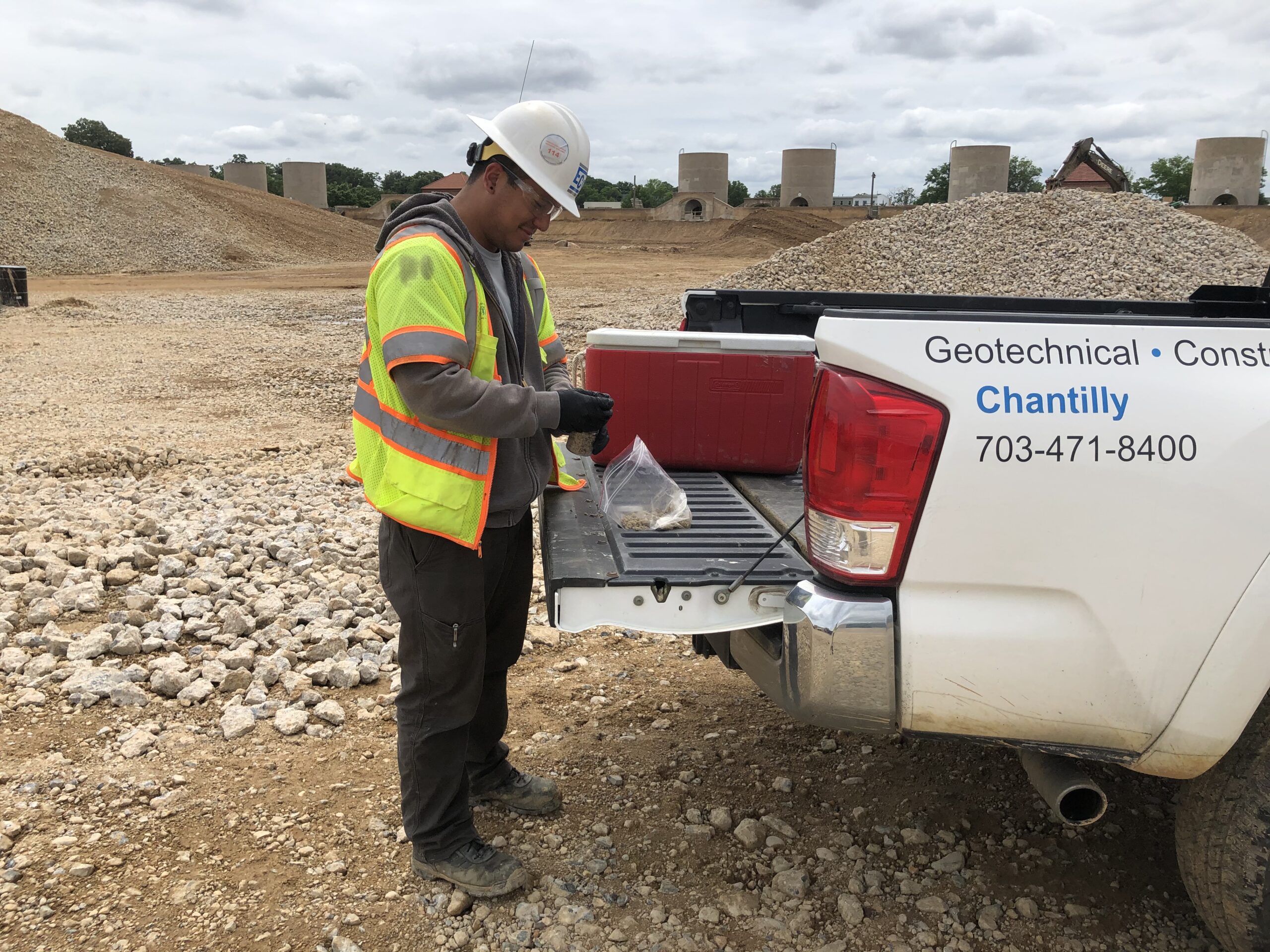 Photo of a man in a white hard hat and neon safety vest standing at the open tailgate of a white truck. He's working with a clear baggie of soil and small hand-held equipment.