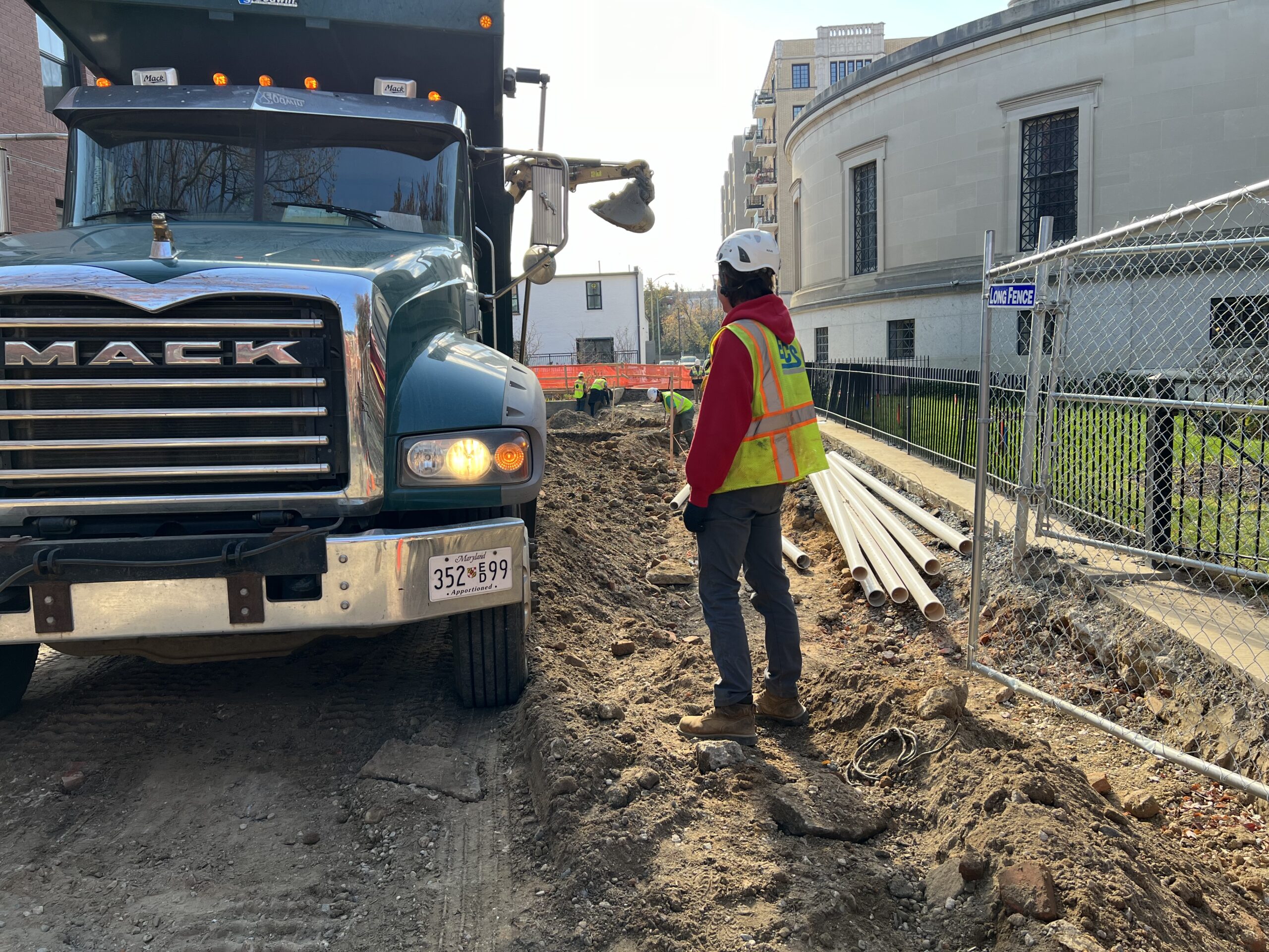 Man in a white hard hat, red hoodie, and neon safety vest stands facing a green Mack truck in a construction area between buildings.