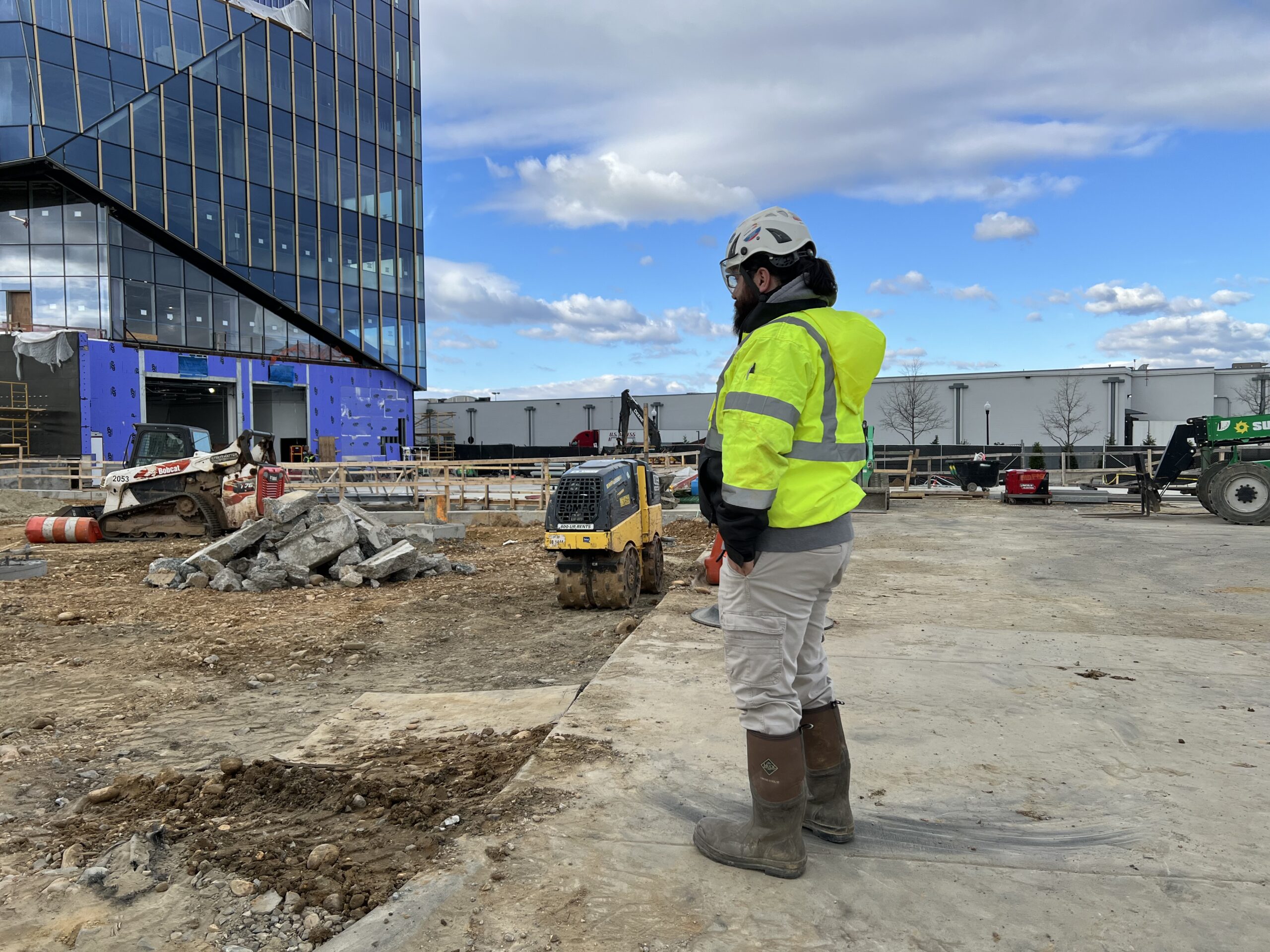 Photo of a man in a white hard hat and neon safety vest stands looking over an excavation site in front of a glass building.