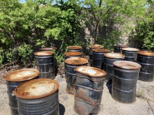 Photo of several rusted and dented oil drums sitting on a concrete pad in front of a fence covered in greenery.