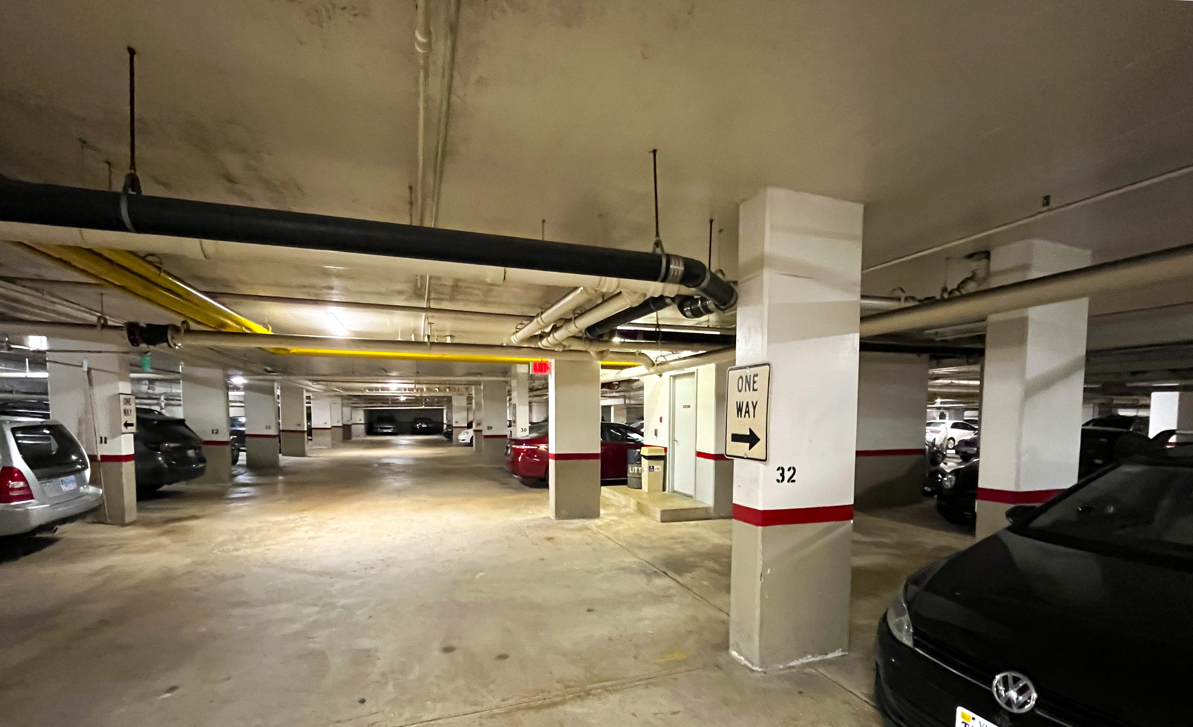 Photo of the interior of an underground parking garage. There are pipes securely mounted to the ceiling with concrete pillars and flooring and cars parked in spaces between the pillars.