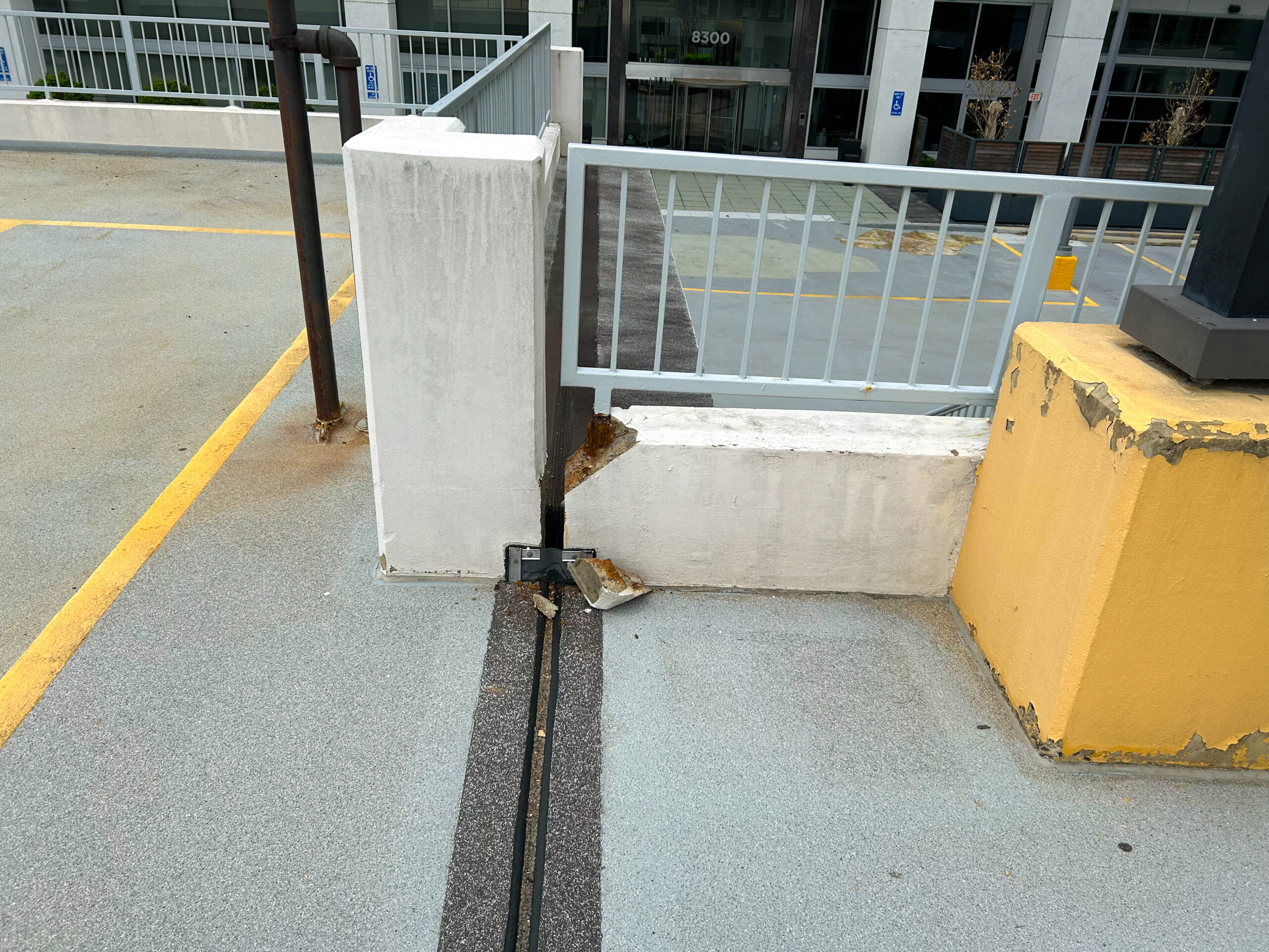 Photo of the top level of a parking structure where portions of the parking garage meet at a metal seam and the concrete wall and bannister show damage. A large chunk of concrete has fallen off the corner of the wall to the floor of the structure.
