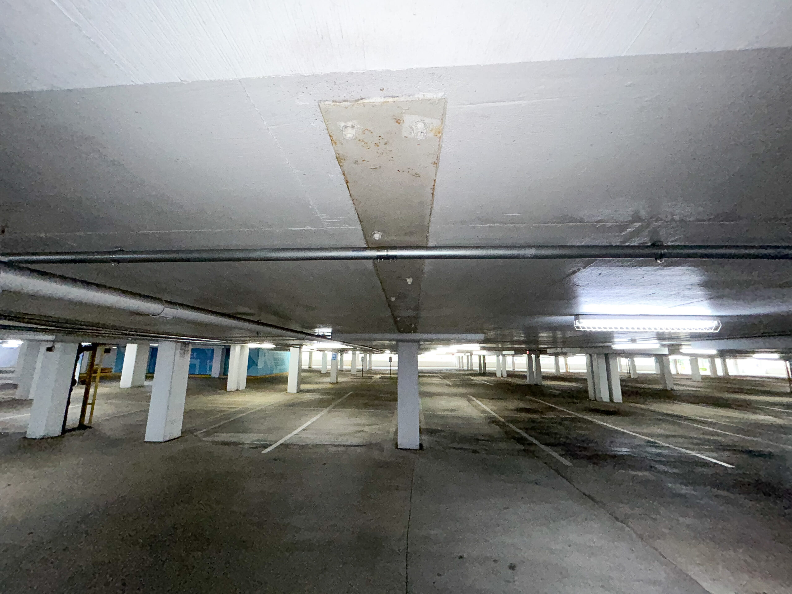 Photo of the inside of a parking structure where the parking garage shows damage along the ceiling in the foreground. In the background there are supporting columns and concrete marked with parking spaces.