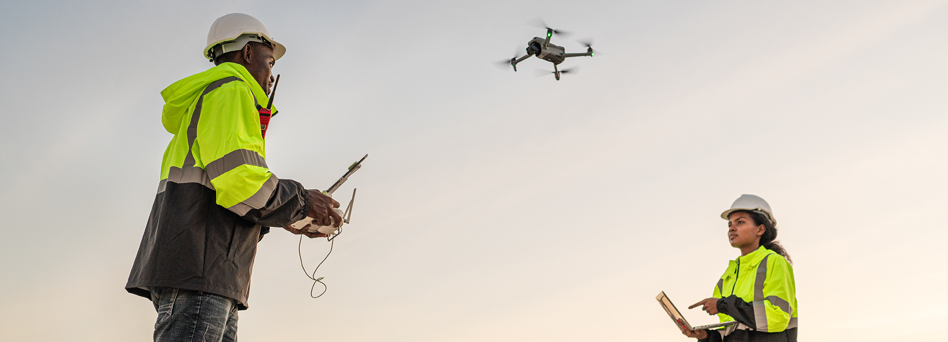Two engineers in high-visibility safety jackets and hard hats operate an unmanned aircraft system at a construction site. One person controls the drone with a remote, while the other monitors data on a tablet as the drone hovers in the air during daylight.