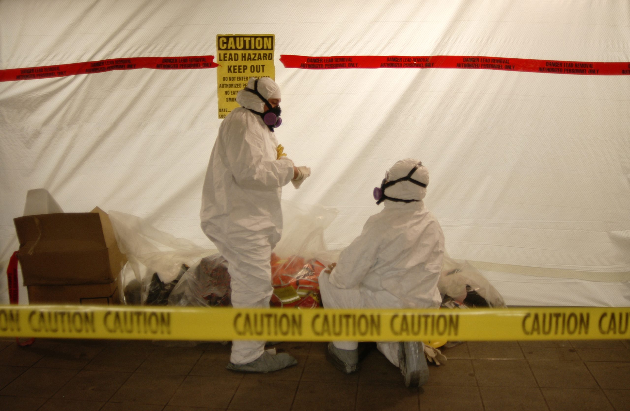 Two workers in full hazmat suits conduct lead paint surveys inside a sealed containment area. The scene is marked with yellow caution tape and a warning sign indicating hazardous material. Debris and sealed waste bags are visible on the floor inside the enclosure.