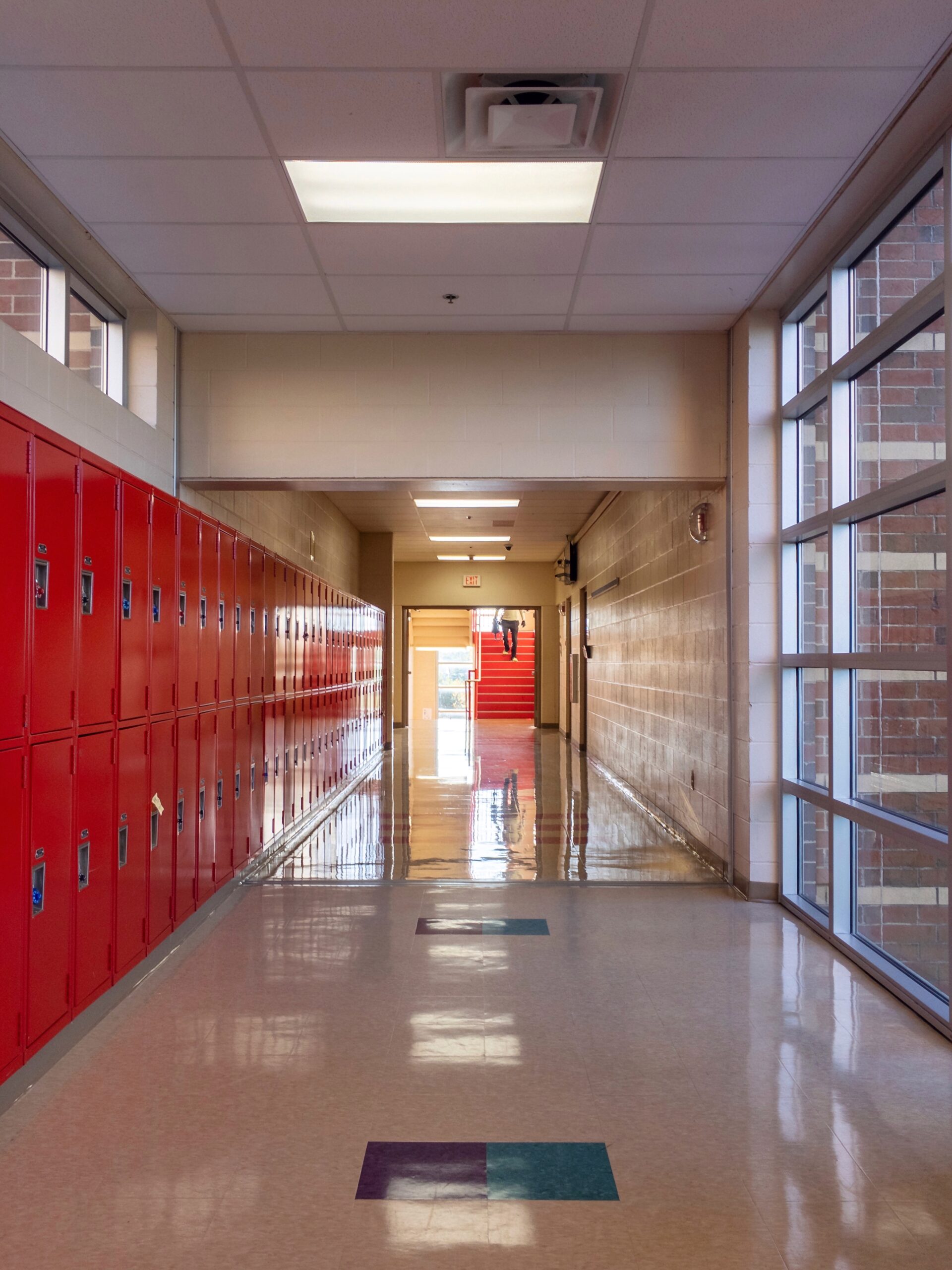 A clean, well-lit school hallway with red lockers and large windows, representing a facility maintained with strict industrial hygiene + hazardous material management protocols and AHERA compliance to ensure student and staff safety.