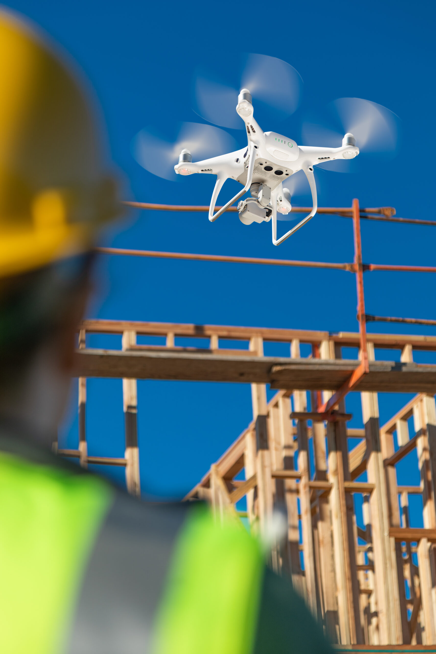 A construction worker wearing a yellow hard hat and neon safety vest watches an unmanned aircraft system (UAS) drone hovering near a wooden building frame under a clear blue sky.